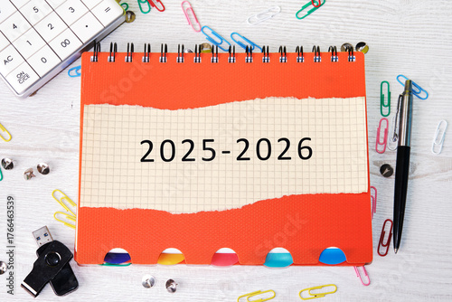 A vibrant orange planner lies open, showing the years 2025-2026. Surrounding it are office supplies like clips and a USB drive, ideal for busy days ahead