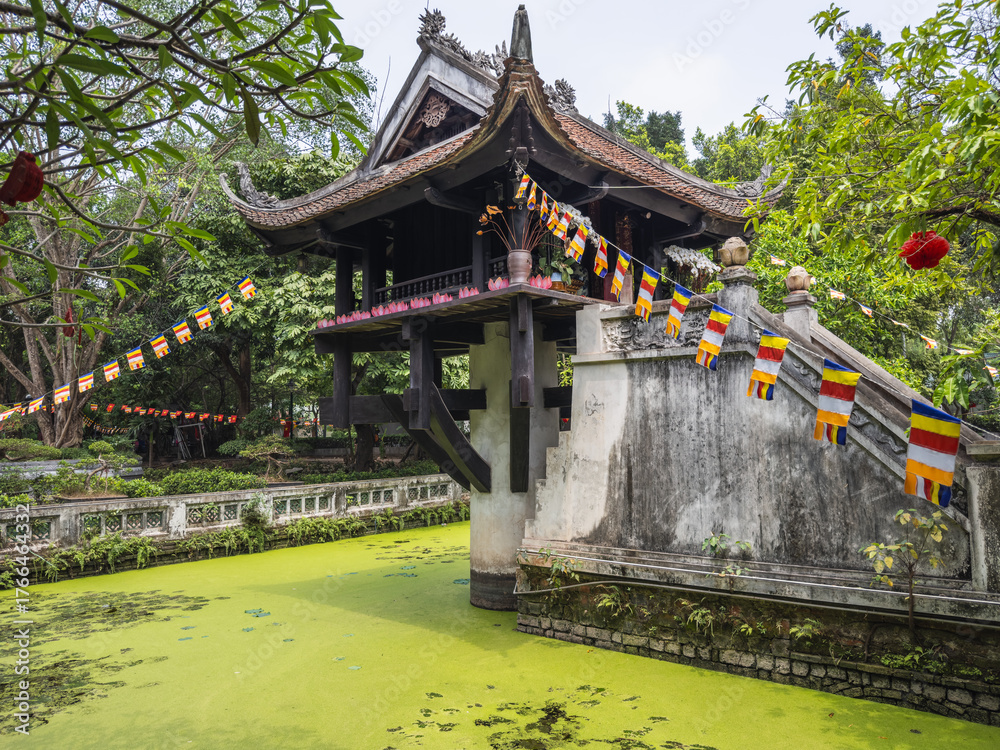 Obraz premium The iconic One Pillar Pagoda in Hanoi, Vietnam, rises elegantly from its lotus pond, adorned with vibrant Buddhist prayer flags amidst lush tropical greenery. A serene cultural landmark.