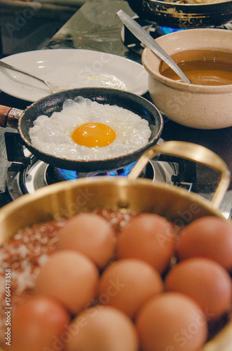 A sunny-side egg cooks in a small cast-iron pan over a blue gas flame. A bowl of whole eggs sits in the foreground at a busy kitchen station. Warm light, shallow depth of field, and natural food styli