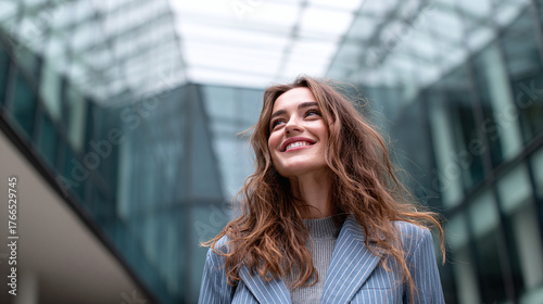 Smiling woman gazes upward amidst city's modern architecture. She wears a striped blazer, radiating joy and optimism in the urban landscape.