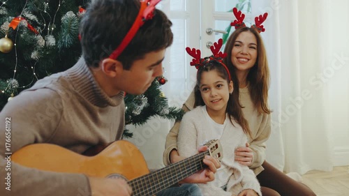 Family enjoys a cozy holiday moment while singing and playing guitar near a Christmas tree decorated with ornaments and lights