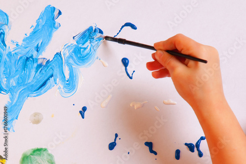 Close-up of child’s hand painting with blue brush on white paper. Kids creativity and art education concept.