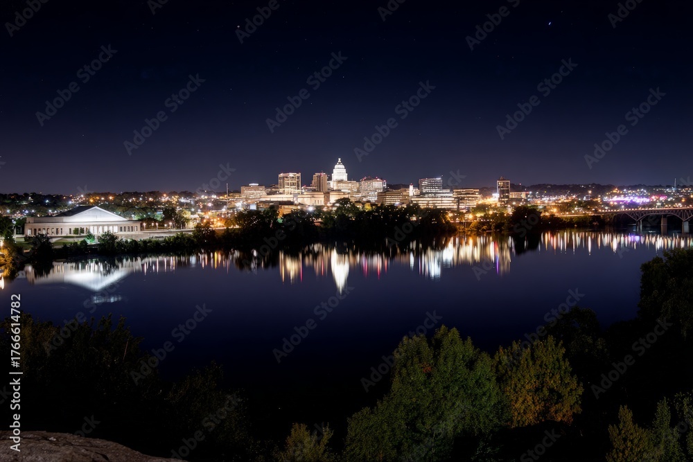 Fototapeta premium City lights glow brightly at night, modern buildings reflect on smooth river surface, dark starry sky