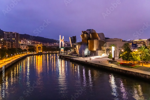 View of the Guggenheim Museum's titanium curves reflecting the river's dark waters under a twilight sky, framed by city lights, Bilbao, Spain.
