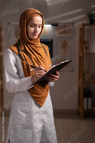 Portrait of Muslim doctor woman wearing hijab writing notes in clipboard in modern office looking at camera working on night shift in hospital. Profession, medicine and healthcare