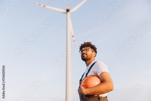 Technician holding helmet looking up at wind turbine in sustainable energy plant