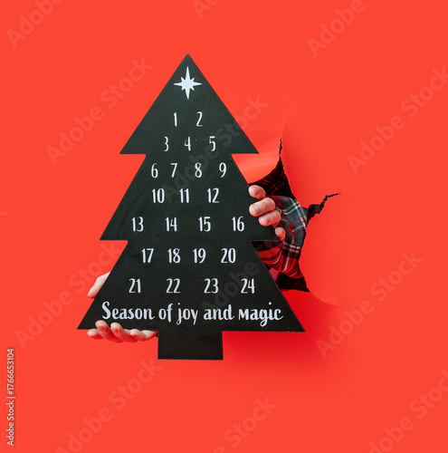 Merry Christmas. person in plaid shirt holds black Christmas tree-shaped advent calendar through torn red paper, marking dates with white pen.