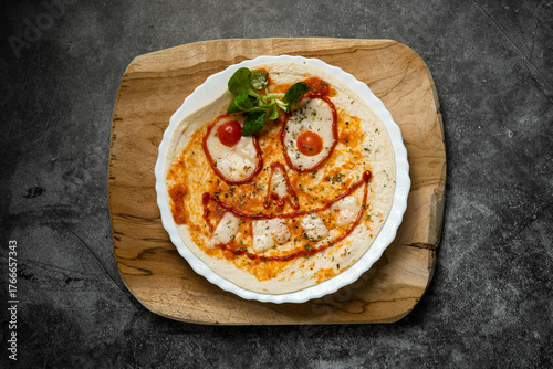 Close-up of Halloween-themed pizza with smiling face made from vegetables and cheese on white plate over wooden board, top view food photography