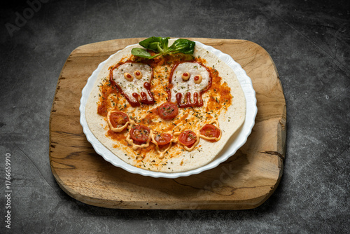 Close-up of Halloween-themed pizza with skull-shaped eyes and tomato mouth on white plate over wooden board, top view food photography for spooky presentation