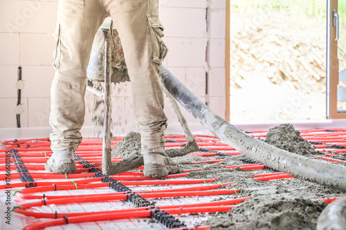 A construction worker efficiently operates a concrete pump to pour concrete onto heated flooring installed with red hoses for a modern building project's thermal system.