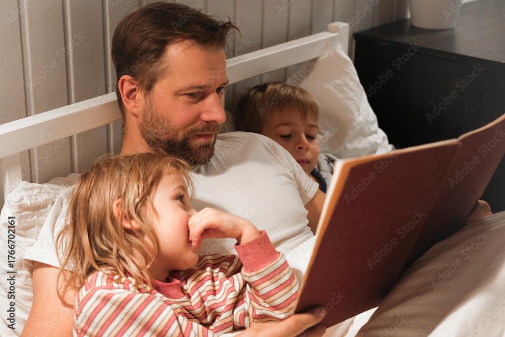 Fototapeta premium Happy mature 40 year old father reading book to his curious children lying in a bed before the sleep. Dad, little daughter and son spending time together. Family read the fairy story. Evening bedroom.