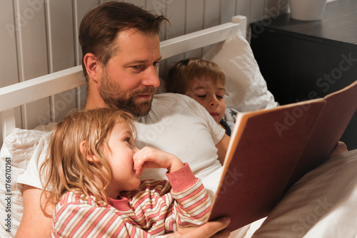 Happy mature 40 year old father reading book to his curious children lying in a bed before the sleep. Dad, little daughter and son spending time together. Family read the fairy story. Evening bedroom.