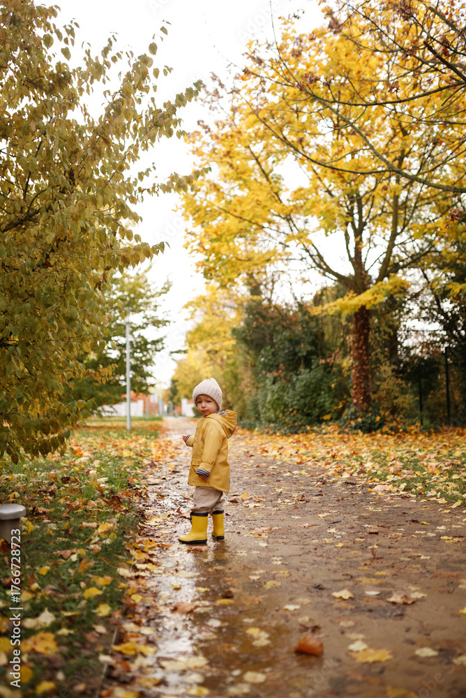 Fototapeta premium Little child wearing yellow raincoat and hat walking through fallen autumn leaves on path outdoors. Cozy fall atmosphere, family walk, childhood exploration and seasonal concept.