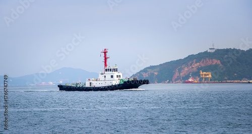 Valokuva Assistance Tug Vessel Cruising Near a Hilly Coastline and Port Facilities