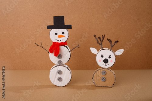 Cardboard snowman and reindeer handmade Christmas crafts with buttons, scarf, hat, and twig antlers on brown background. DIY winter decoration project. Studio composition for greeting card, poster
