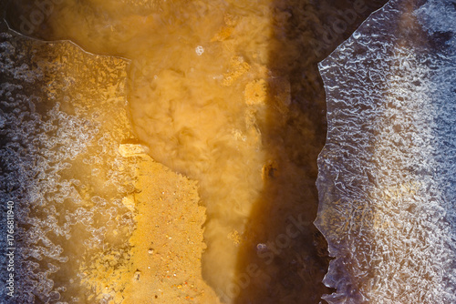 Dirty silt and sand rising from a shallow stream bed, with fragmented ice shards floating. March thaw, abstract nature texture