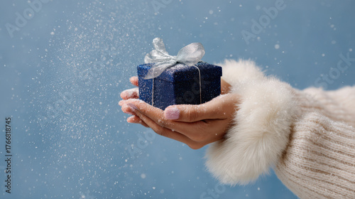 Hand holding a blue gift box with silver ribbon, surrounded by falling snowflakes for festive Christmas celebration with copy space