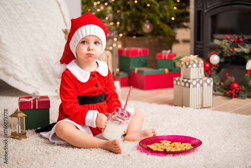 Kid in santa costume with milk and cookies on fluffy rug, surrounded by christmas tree, gifts, and fireplace.