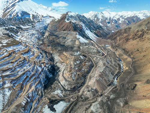 High aerial view of an enormous industrial mining complex and terraced quarry carved into a rugged, snow-dusted mountain valley floor beneath high snowy peaks