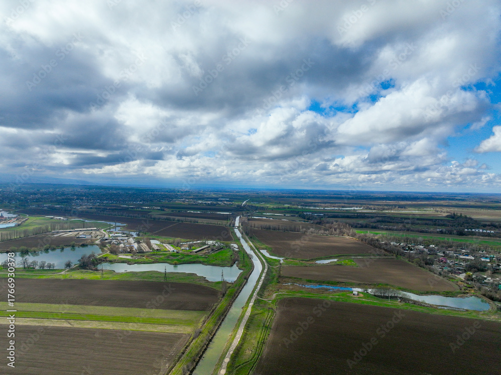 Fototapeta premium Wide aerial view of a straight canal and several interconnected water bodies running through dark brown and green agricultural fields in a flat valley