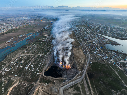 High vertical aerial shot of an intense column of fire and thick smoke rising from a landfill situated between residential neighborhoods and a river