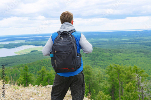 A man in a blue tank top and gray turtleneck stands on a mountain top with a backpack.