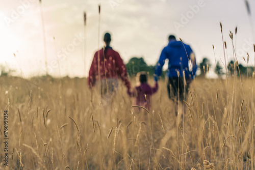 Blurred family of three holding hands, walking together through a golden wheat field towards the sunset.