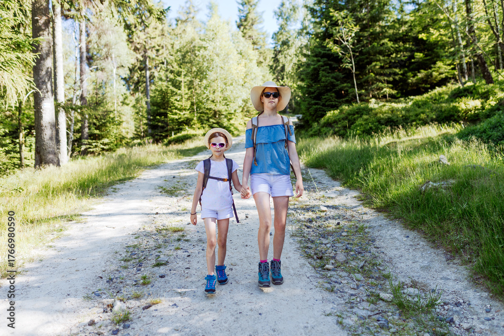 Fototapeta premium Two sisters hikers with backpacks, hats and sunglasses walking together along a sunny forest trail, summer outdoor adventure, family bonding and active lifestyle in nature scene