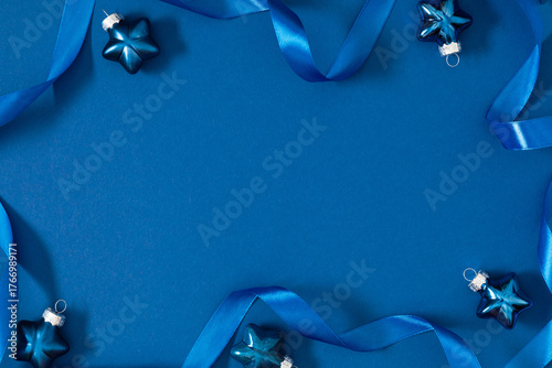 Blue ribbon and star-shaped Christmas ornaments on blue background. Minimalist festive composition with copy space. Elegant winter layout for greeting cards and holiday design