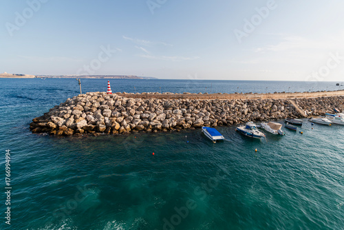 Papier peint A rocky breakwater with a striped lighthouse extends into the vibrant blue sea, protecting several moored boats under a clear sky