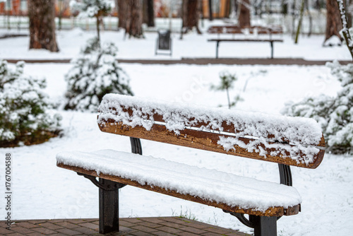 A wooden park bench is completely covered in fresh, white snow after a snowfall. The quiet winter scene highlights the serene beauty of a deserted park during the cold season.