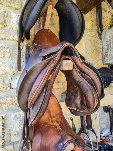 Fotografi Rustic collection of worn, brown leather saddles and black tack hanging on a woo