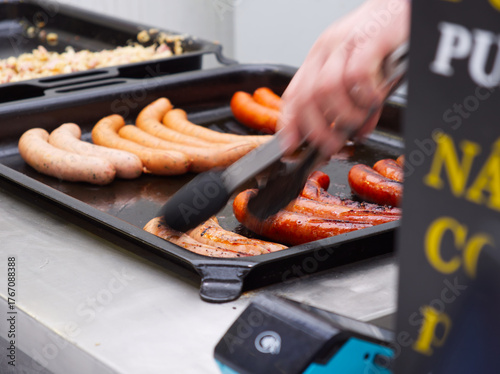 Quadro su tela Hand using tongs to cook various sausages on black electric griddle pan in kitch