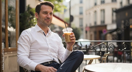 A smiling man in a white shirt enjoys an iced drink at an outdoor cafe on a sunny day in a European city.
