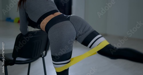 focused athletic woman executes a bent over glute kickback while holding onto a chair for support in a bright fitness studio. Her workout emphasizes glute strength and stability.
