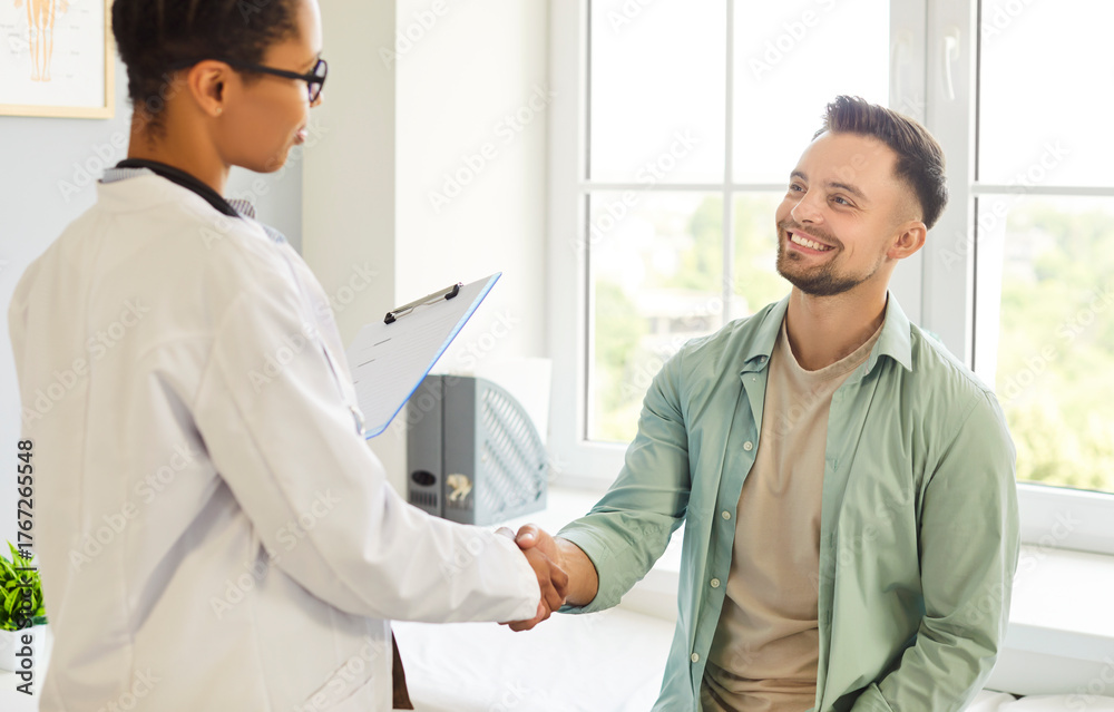 Fototapeta premium Smiling friendly happy female doctor therapist in white uniform shaking hands with a young patient man standing in office after medical examination in clinic. Medicine and health care concept.