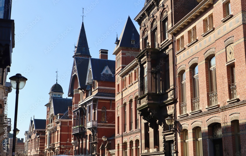 Obraz premium Street view of historic eclectic townhouses in Antwerp, featuring red brick facades, dark slate roofs, and turrets against a clear blue sky.