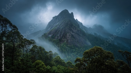 A towering mountain rises above dense green forests shrouded in mist and clouds during early morning hours.