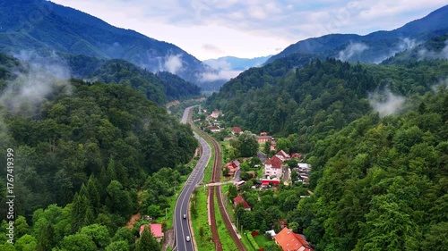 Mountain valley with road and village surrounded by forest, Romania. Aerial panorama of a road, railway, and small village nestled between green forested mountains in the Carpathians, Romania.