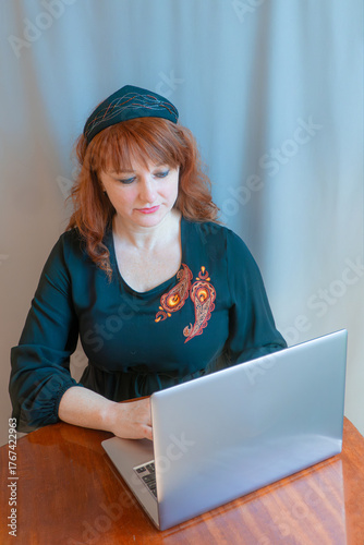 A beautiful young woman is working or playing or shopping on a laptop in a stylish modern dress and a hair band in the shape of the Russian national kokoshnik headdress