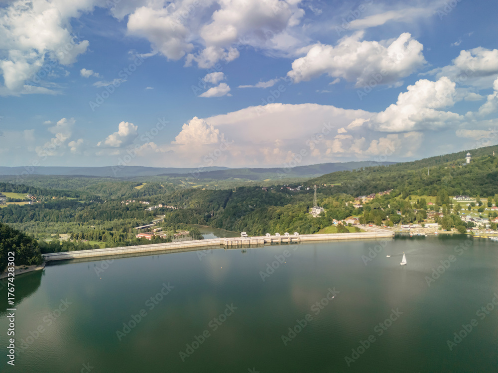 Obraz premium Aerial view of Solina Dam, Poland, showcasing the serene lake surrounded by lush green hills under a bright blue sky with fluffy clouds. Ideal for nature and travel enthusiasts.