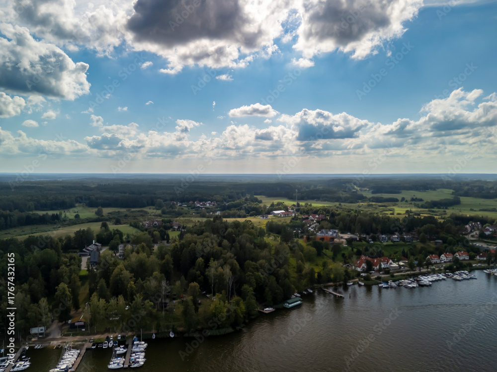 Obraz premium Aerial view of Mikolajki, Poland, showcasing a vibrant landscape with lush green trees, a calm lake, and numerous boats docked along the shoreline under a vast sky with clouds.