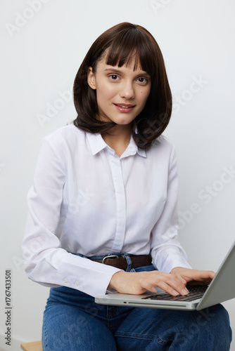 A young woman sits with a laptop on her lap in a bright, clean space. She smiles softly, wearing a white shirt and jeans, ready for online tasks and everyday work from home.