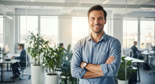 Confident businessman with arms crossed smiling in a modern, sunlit office environment with colleagues working in the background.