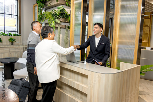 Happy senior Asian couple checking in at hotel on holiday vacation with young male receptionist. Receptionist man in suit shaking hands with Elderly traveler customer at hotel reception counter desk.