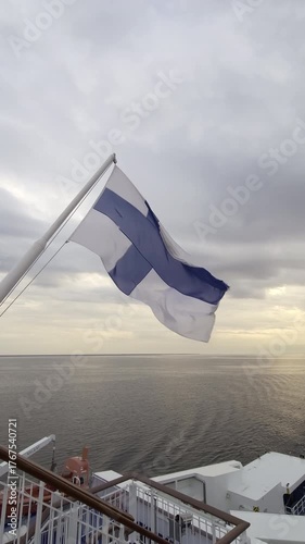 Waving flag of Finland on the stern of a large ferry sailing on the Baltic Sea, with the ship's wake visible on the water and a cloudy sky during a beautiful golden hour sunset