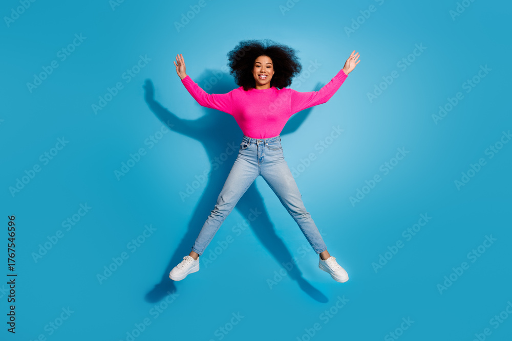 Obraz premium Cheerful young woman in a bright pink top smiling and jumping against a blue backdrop, expressing joy and vibrance