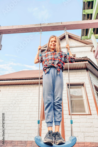 A young Caucasian girl with long blonde hair swings joyfully on a playground swing. She wears a plaid shirt and gray pants, with a house in the background.