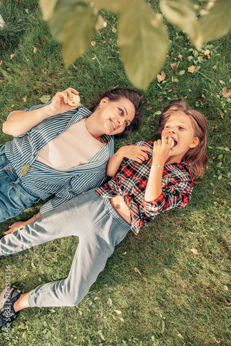Two young girls lying on grass, enjoying snacks. One has dark hair and wears a striped shirt, the other has light brown hair in a plaid shirt. They share a joyful moment.