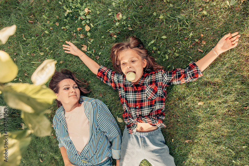 Two young girls lying on green grass. One girl has long brown hair and wears a plaid shirt. The other has medium-length brown hair and wears a denim jacket. They enjoy a sunny day.